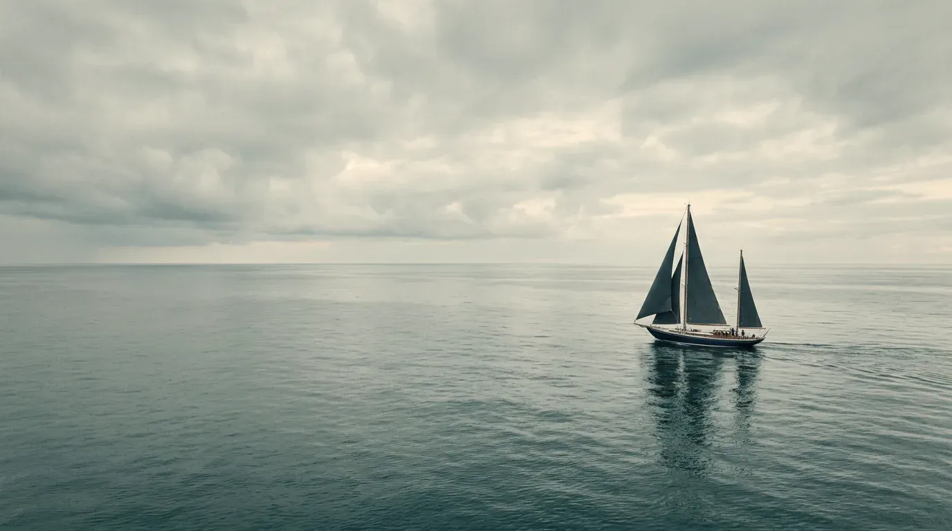 A classic two-masted yacht under sail on calm open water, beneath a wide overcast sky