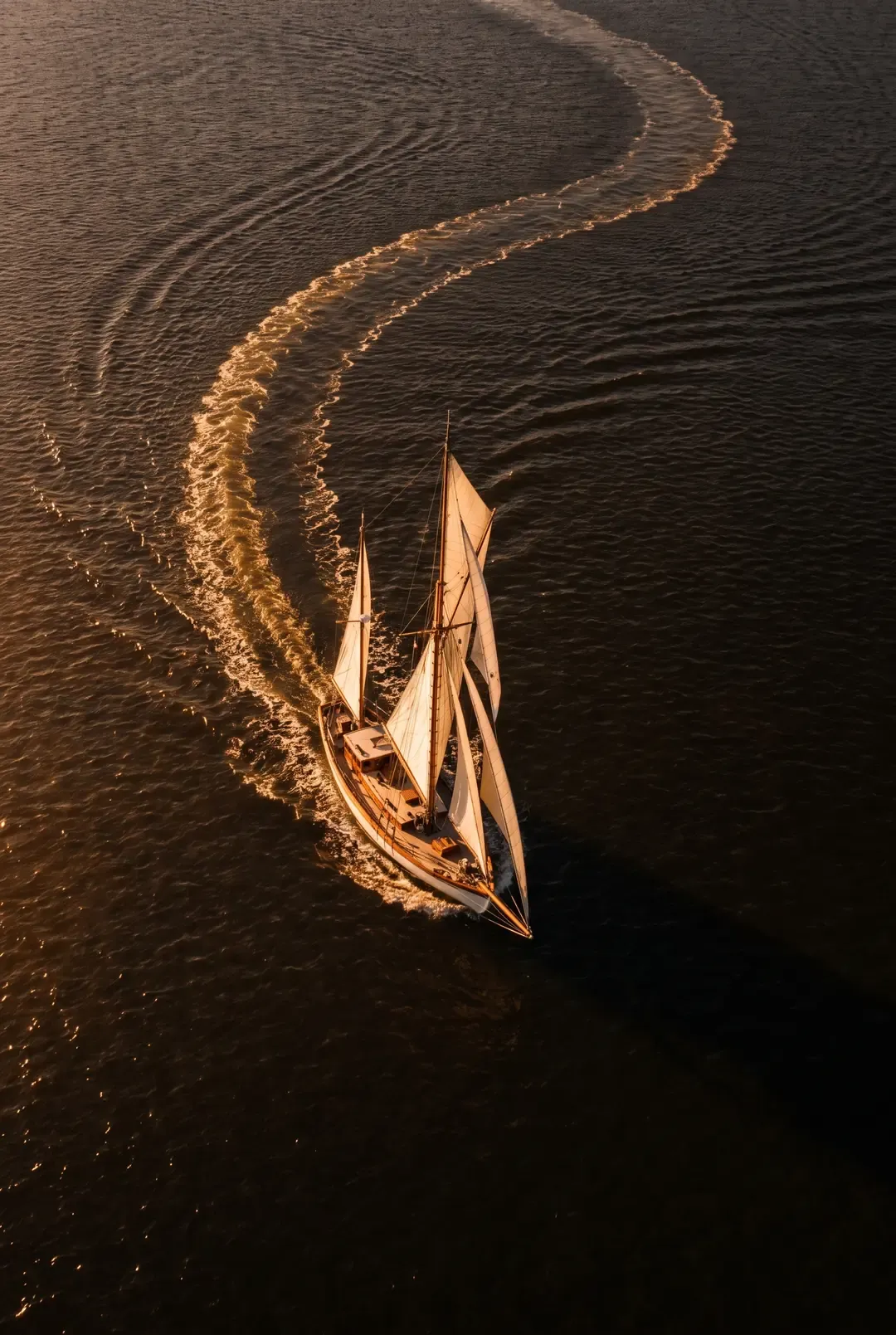 A classic yacht under sail at dusk, its wake curving behind on bronze water